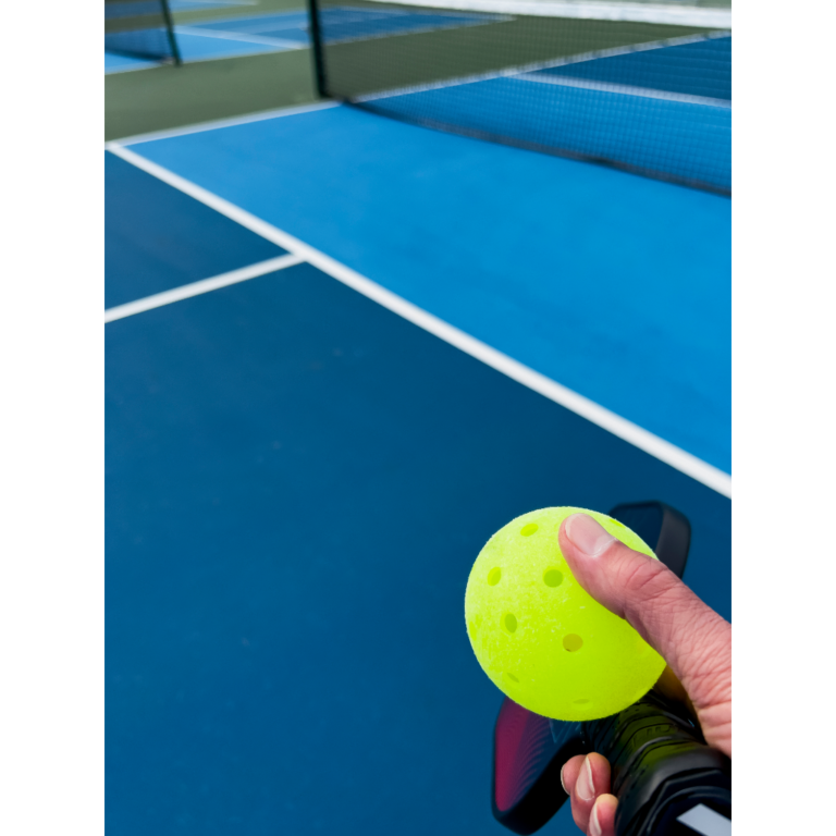 A pickleball player holding a pickleball ball and paddle on a pickleball court.